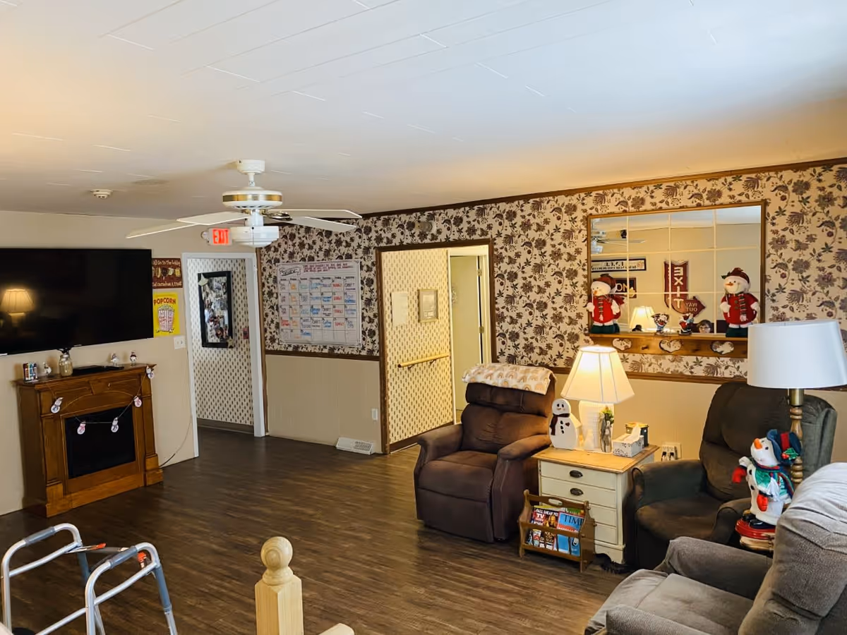 Community living room with recliners, a wall-mounted TV above a faux fireplace, side tables with lamps and holiday decorations.