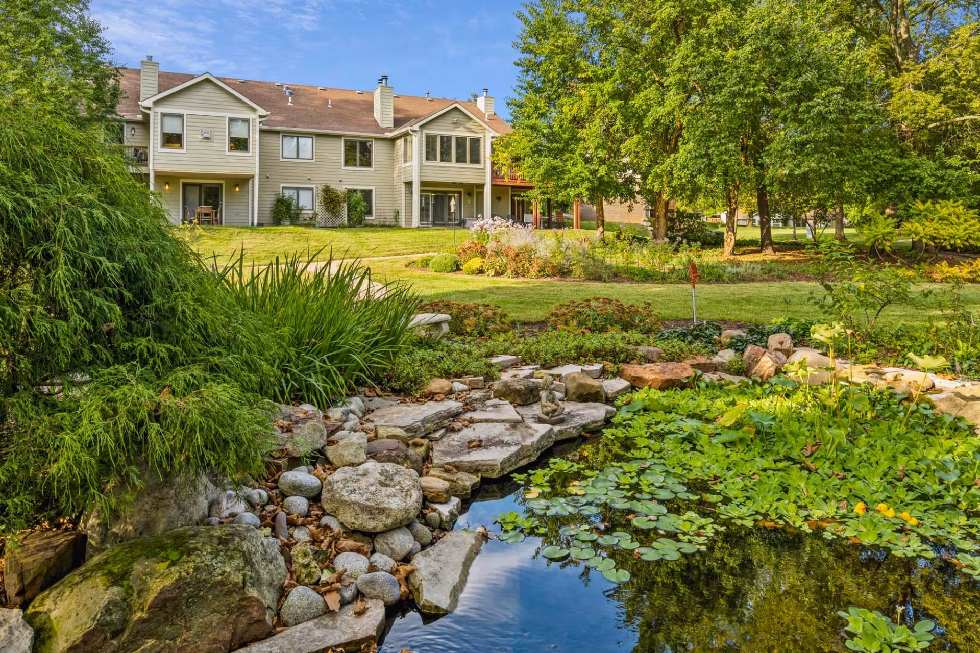 A landscaped garden area with a small pond filled with lily pads and surrounded by rocks and greenery. In the background, there is a two-story residential building with beige siding and multiple windows, set against a blue sky with some clouds. Trees and shrubs are also visible around the garden.