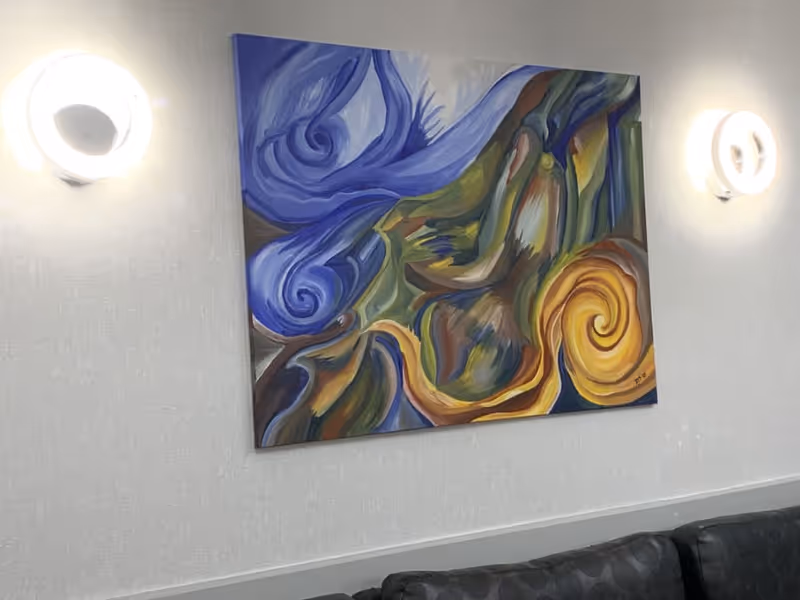 Abstract colorful painting with swirling patterns in blue, yellow, green, and brown hues hanging on a light-colored wall between two circular wall lights above a dark patterned couch.