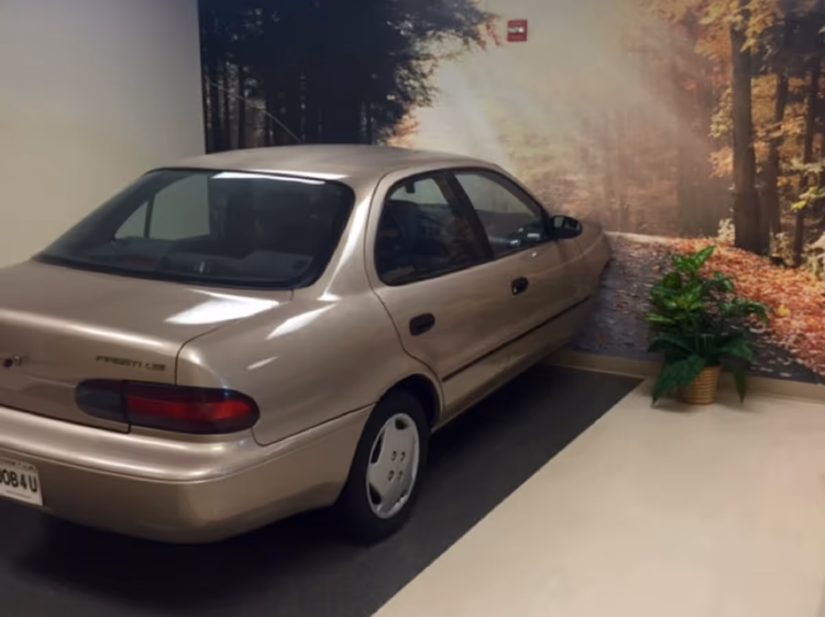A beige sedan car parked indoors next to a wall mural depicting a forest pathway with autumn leaves. A green potted plant is placed near the wall on the right side of the car.