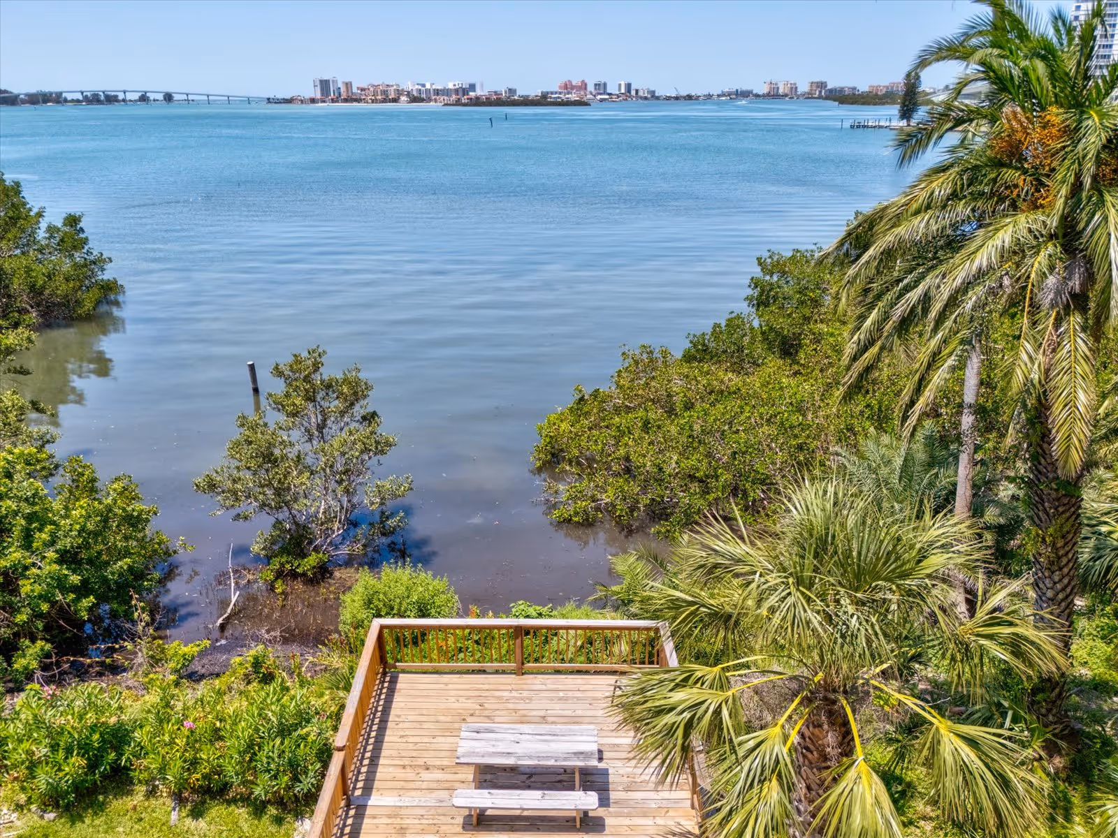 View of a wooden deck with a picnic table overlooking a calm body of water surrounded by lush greenery and palm trees, with a city skyline and bridge visible in the distance under a clear blue sky.