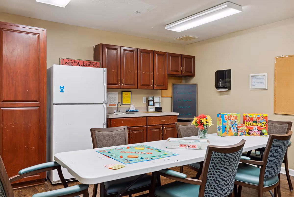 A room with a white table and four chairs, featuring board games like Monopoly, KerPlunk, Uno, and Hedbanz on the table. Behind the table is a kitchenette area with a white refrigerator, wooden cabinets, a sink, and a small chalkboard on the wall. A vase with colorful flowers is placed on the table.