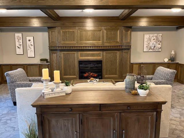 A cozy living room with a wooden cabinet in the foreground topped with decorative items including candles, a small plant, and a sign that reads 'this feels like home'. Behind the cabinet is a beige sofa facing a wooden fireplace with a built-in electric fire. Two patterned armchairs flank the sofa. The walls are decorated with framed floral artwork, and the ceiling features exposed wooden beams.