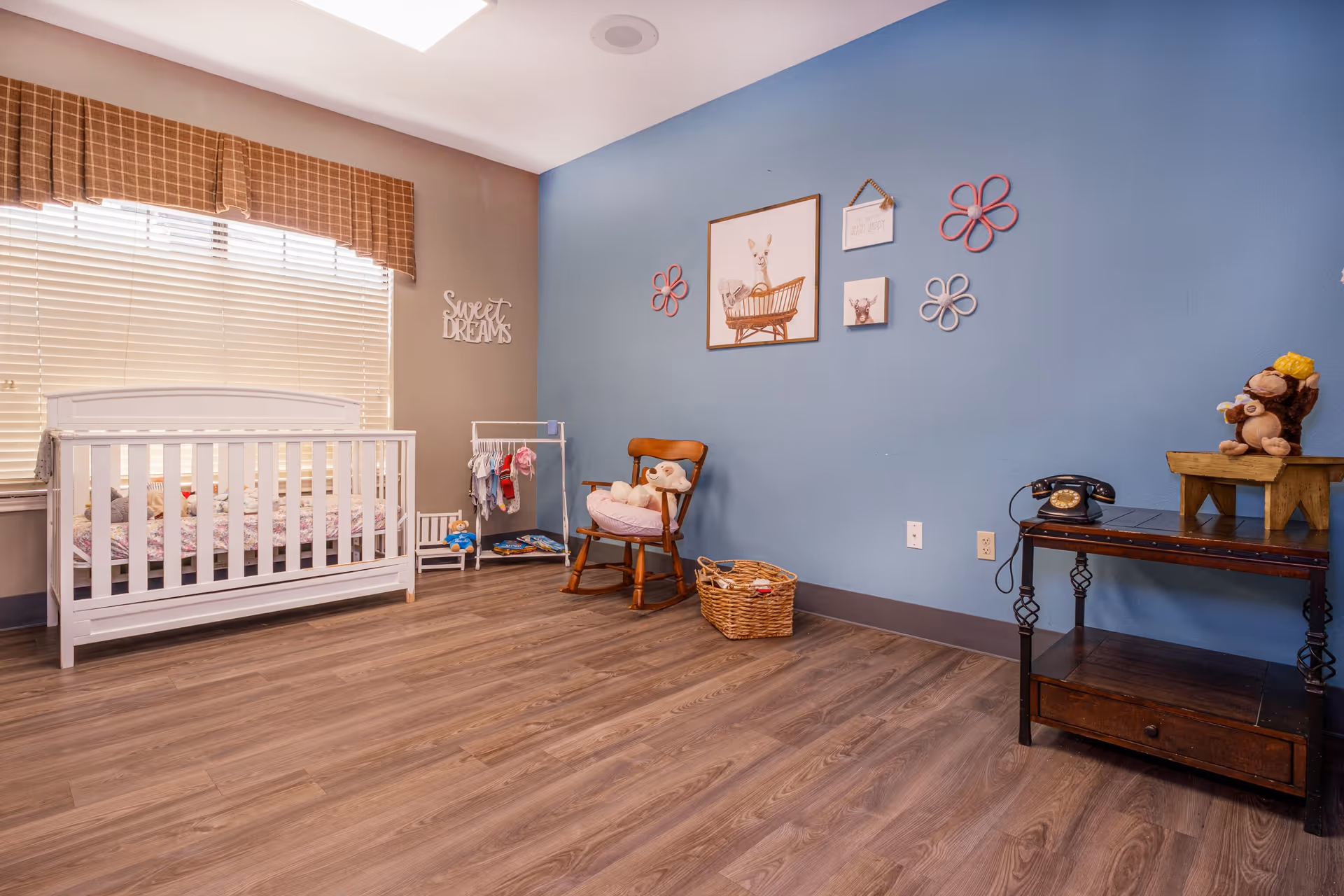 A cozy nursery room with a white crib near a window with blinds and a brown valance. The walls are painted blue and beige, decorated with framed animal pictures and flower-shaped wall hangings. There is a wooden rocking chair with a teddy bear, a small clothes rack with baby clothes, a wicker basket on the floor, and a dark wooden table holding a vintage black rotary phone and a stuffed monkey toy on a small wooden stool.