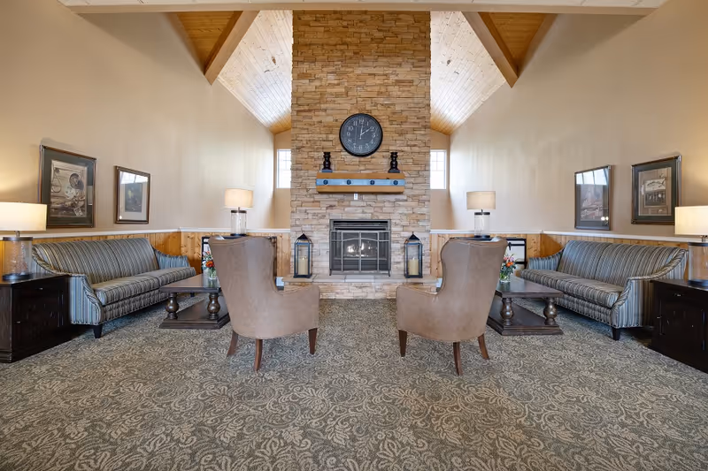 A cozy living room area with two striped sofas facing each other, two beige armchairs facing a stone fireplace with a clock above it. The room has a patterned carpet, wooden ceiling beams, and lamps on side tables next to the sofas.