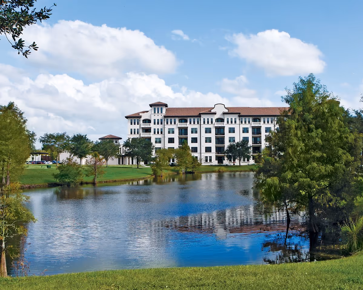 A large white multi-story building with a red-tiled roof is situated behind a pond surrounded by green grass and trees under a partly cloudy blue sky.