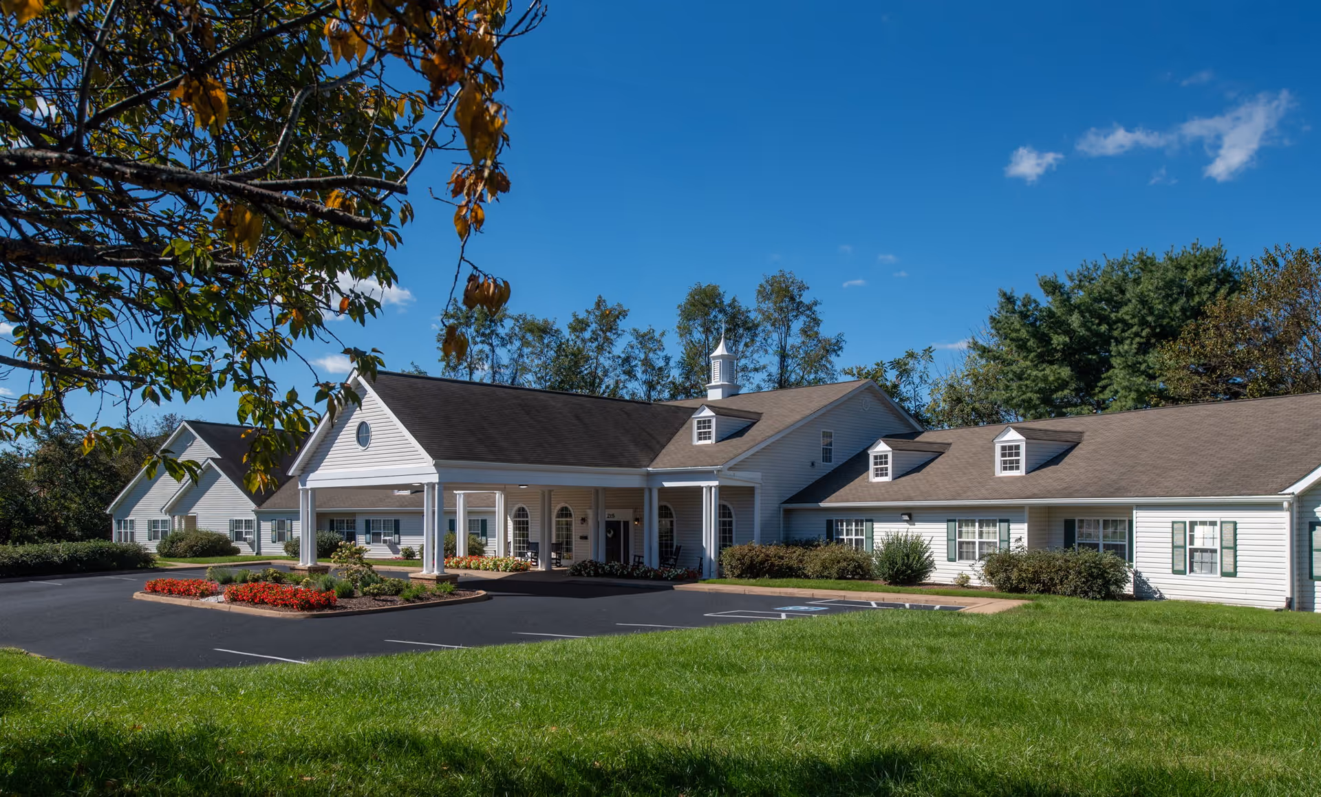 Front exterior of a single-story white senior living facility with a covered entrance, landscaped grounds, and parking area under a blue sky.