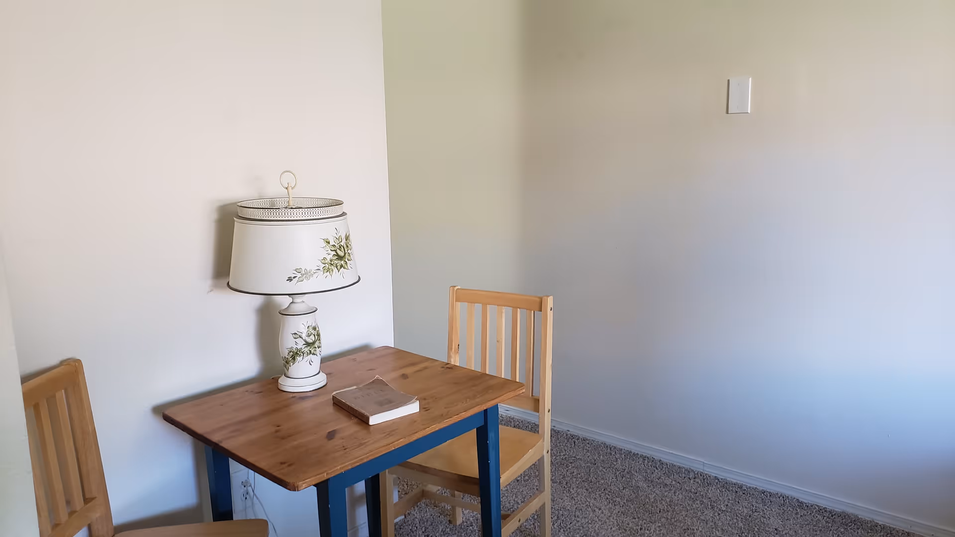 A small wooden table with blue legs and two wooden chairs in a corner of a room with light-colored walls and carpeted floor. On the table, there is a decorative white lamp with floral patterns and a book.