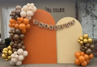 A decorative autumn-themed display outside a building with the address 180 Starr. The display features two large curved panels in orange and cream colors, with a banner across them spelling THANKFUL. There are clusters of balloons in shades of brown, orange, cream, and gold arranged on the left and right sides of the panels.