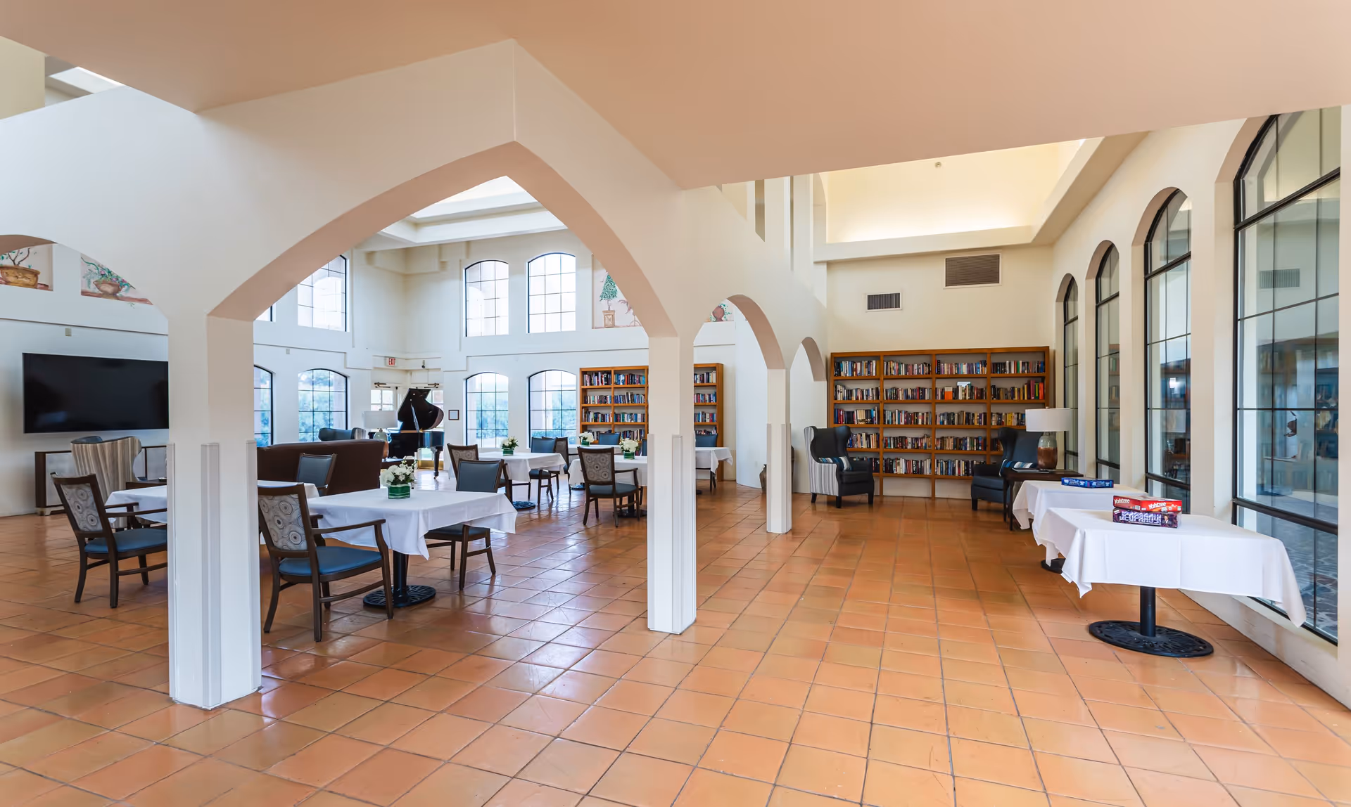 Spacious senior living common area with terracotta tiled floor, white walls, and large arched windows letting in natural light. The room features several tables with white tablecloths and chairs, bookshelves filled with books, two armchairs, and a grand piano near the windows. There are also two tables with board games on them near the windows.