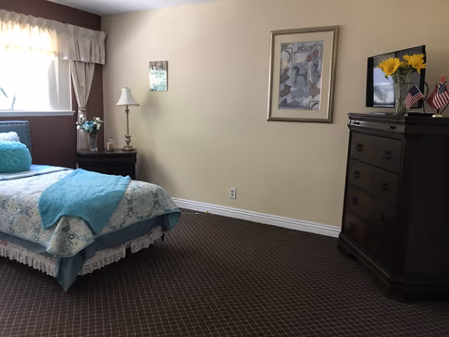 A neatly arranged bedroom with a single bed covered in a light blue and white patterned bedspread and a turquoise throw blanket. There is a nightstand with a lamp and a vase of flowers next to the bed. A framed abstract painting hangs on the beige wall above a dark wooden dresser with a TV on top, along with a vase of yellow flowers and small flags. The room has a window with sheer curtains allowing natural light to enter.