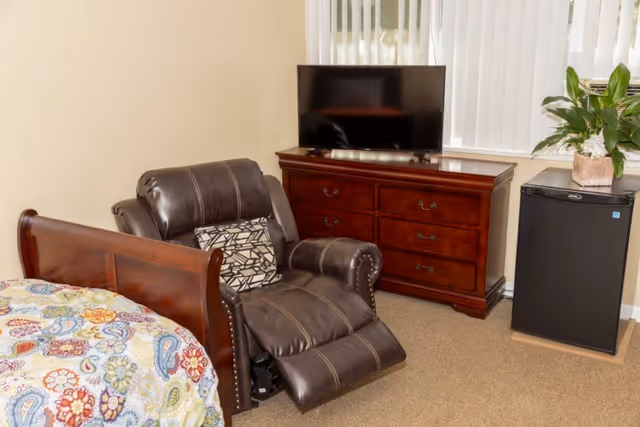 A bedroom with a leather recliner beside a wooden bed, a dresser with a TV, and a mini refrigerator with a plant on top.