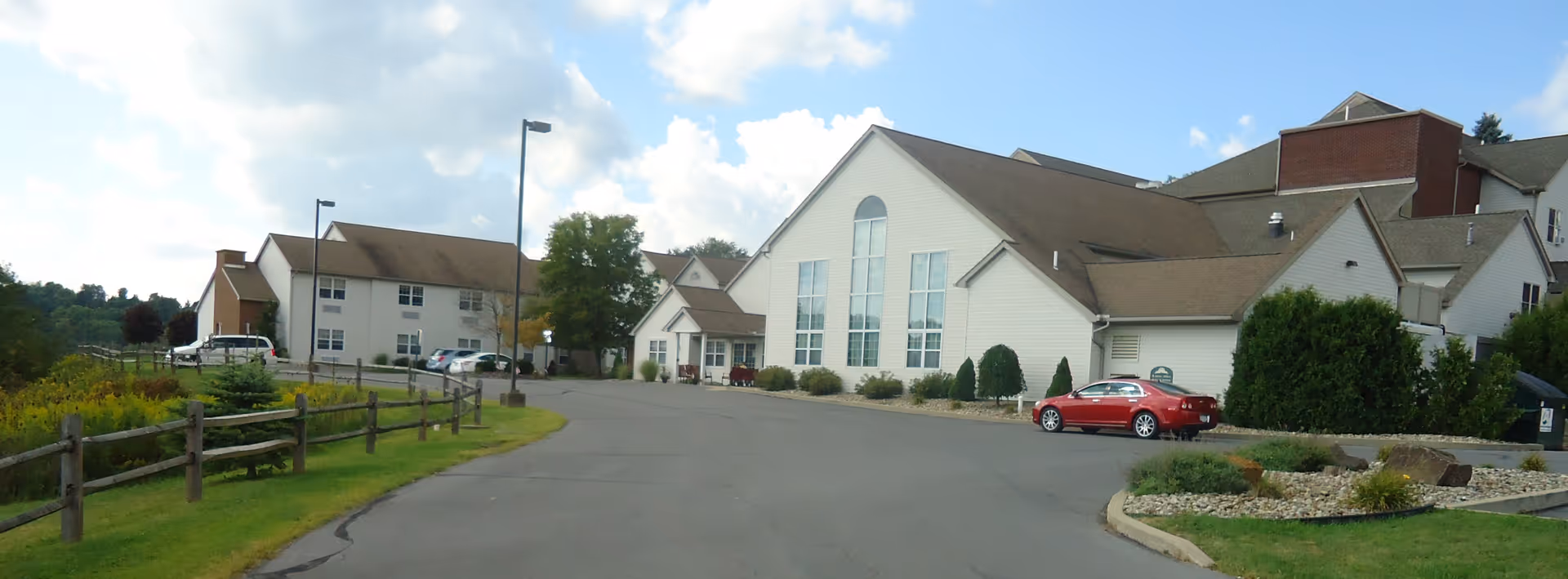 Exterior view of DuBois Village Personal Care Community showing a large building with white siding and multiple windows under a partly cloudy sky. There is a paved driveway with a red car parked near the entrance, surrounded by green grass, bushes, and a wooden fence.