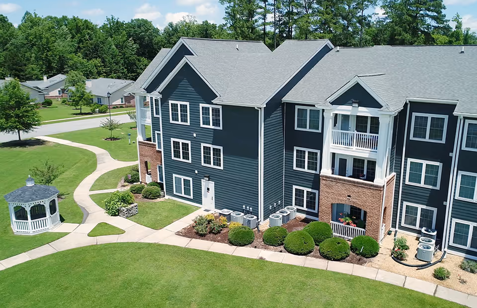 Exterior view of a multi-story residential building with dark siding and brick accents, surrounded by well-maintained green lawns, shrubs, and a white gazebo. Paved walkways curve through the landscaped grounds under a partly cloudy sky.