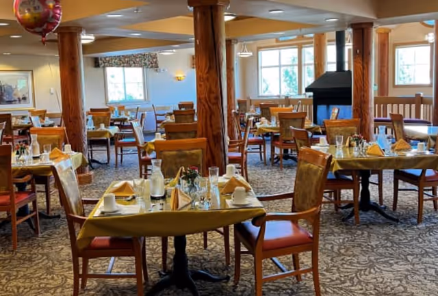 A spacious dining room with multiple tables set for a meal, featuring yellow tablecloths, folded napkins, cups, glasses, and small flower arrangements. The room has wooden pillars, large windows letting in natural light, and a carpeted floor with a patterned design.