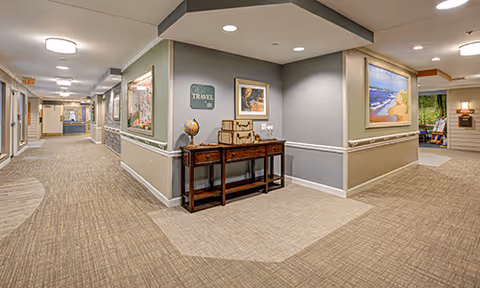 A well-lit hallway in a senior living facility with beige carpet and light gray walls. The hallway features a wooden console table with decorative items including a globe and stacked suitcases. Several framed pictures and artwork hang on the walls. The corridor extends into other rooms and areas, with a reception desk visible in the distance.