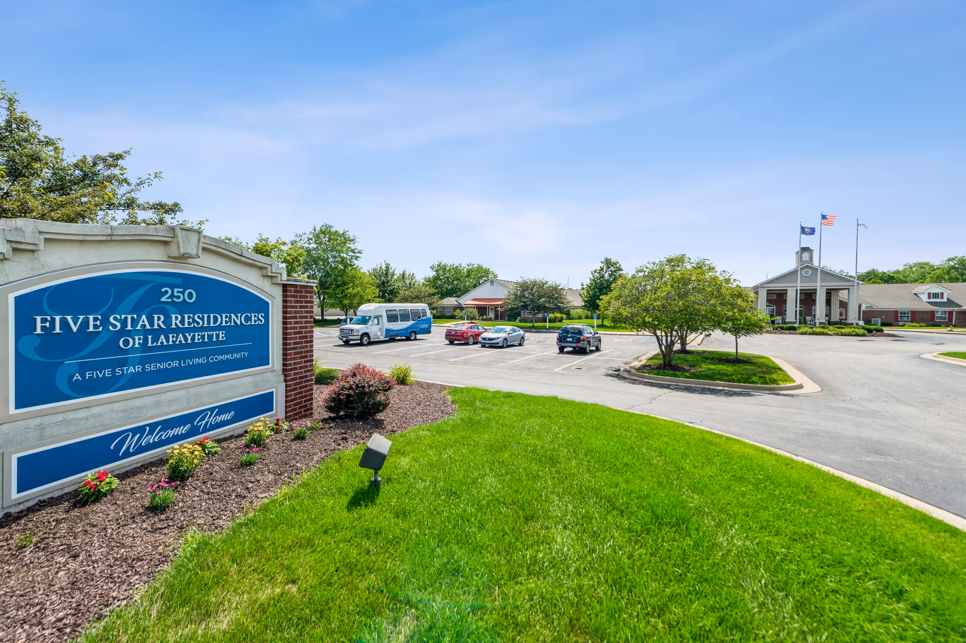 Entrance sign for Five Star Residences of Lafayette beside a driveway and parking lot with the facility building and flags in the background under a blue sky.
