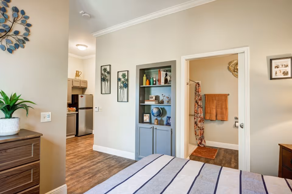 View of a bedroom with a bed in the foreground, a wooden dresser with a potted plant on the left, and an open doorway leading to a bathroom with a shower curtain and towels. To the left of the bathroom is a small kitchen area with a refrigerator and cabinets. The walls are light beige, and there is a built-in shelving unit with decorative items between the bedroom and bathroom.