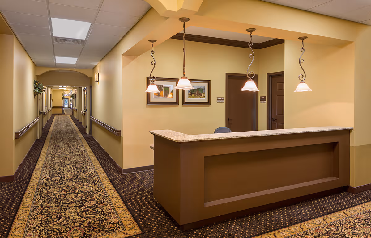 Interior view of a senior living facility hallway with a reception desk on the right. The hallway has patterned carpet and beige walls with handrails. Three pendant lights hang above the reception desk, and framed artwork is displayed on the wall behind it. Two closed doors are visible behind the desk.