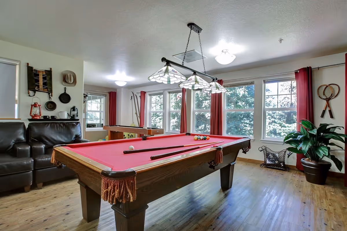 A bright game room with a red felt pool table in the center, two black leather chairs to the left, large windows with red curtains letting in natural light, a potted plant on the right, and various decorative items on the walls including a hat, pans, and tennis rackets.