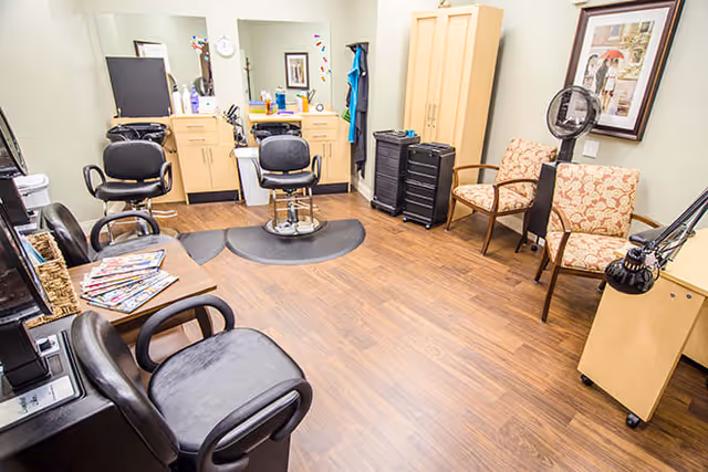 Interior view of a salon area in a senior living facility with two black salon chairs in front of mirrors and wooden cabinets. There are additional black chairs and a small table with magazines, two patterned armchairs, a standing fan, and framed artwork on the wall. The floor is wooden.