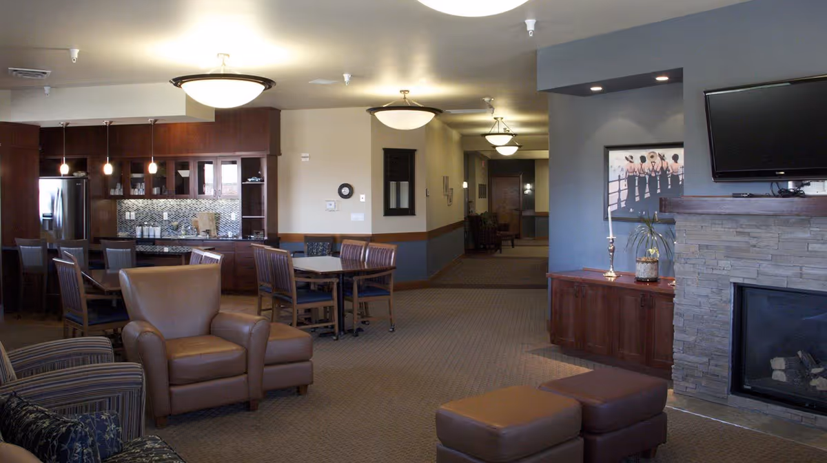 A spacious common area in a senior living facility featuring a seating area with armchairs and ottomans, a stone fireplace with a mounted flat-screen TV above it, a wooden cabinet with decorative items, and a kitchen area with dark wood cabinets, a refrigerator, and a dining table with chairs. The room is well-lit with ceiling lights and pendant lights over the kitchen counter.