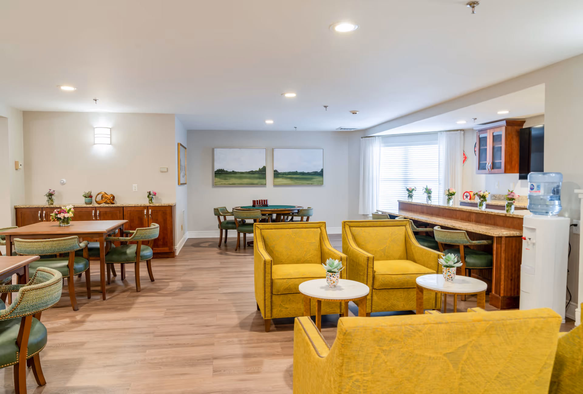 Bright communal lounge and dining area with yellow armchairs, wooden tables, and a service counter in a senior living facility.