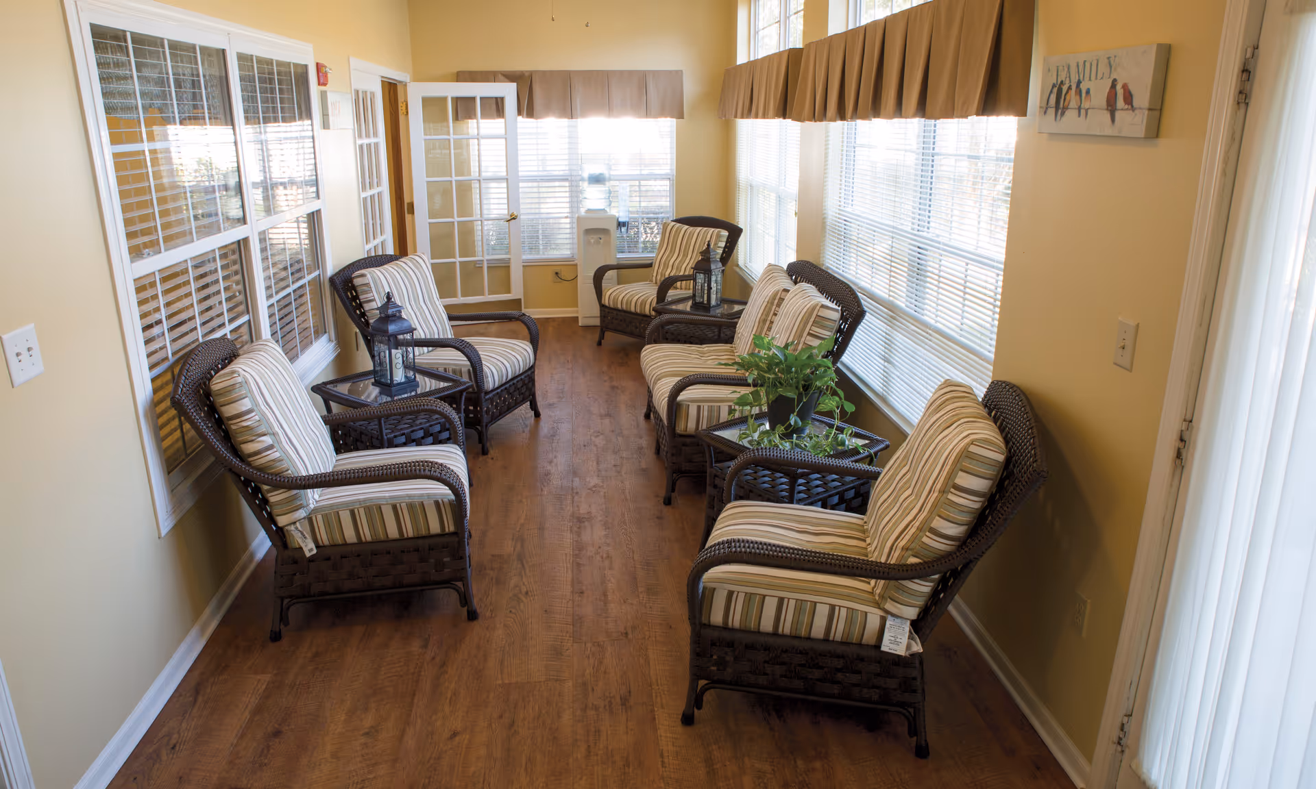 A sunlit sitting area with six cushioned wicker chairs arranged along two walls, each paired with a small glass-top wicker table. The room has large windows with blinds and valances, a wooden floor, and light yellow walls. A water cooler is visible at the far end near a glass-paneled door, and a wall decoration with birds and the word 'FAMILY' hangs on the right wall.