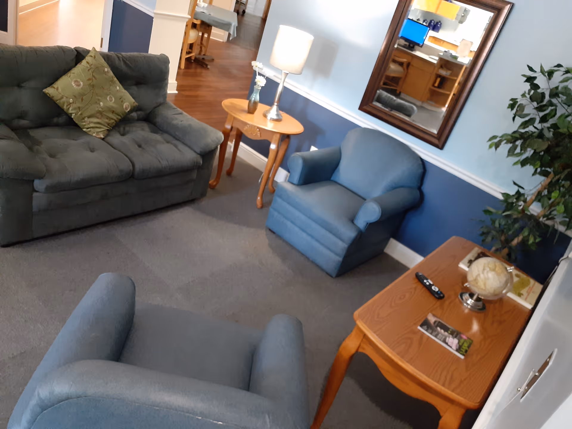 A cozy living room area with a green cushioned loveseat with a decorative pillow, two blue armchairs, a wooden side table with a lamp and small vase, a wooden coffee table with a globe, remote control, and a magazine. A large mirror hangs on the blue and white wall, and a leafy green plant is in the corner. The floor is carpeted and the hallway with wooden flooring is visible in the background.