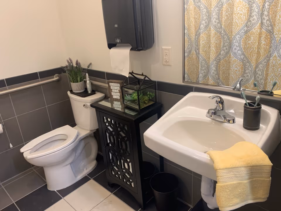 A clean and well-decorated bathroom featuring a white toilet with a grab bar on the wall, a black cabinet with decorative cutouts holding a small plant and a framed quote, a white sink with a chrome faucet and a yellow towel draped over the edge, a toothbrush holder with three toothbrushes, and a large mirror reflecting a patterned shower curtain.