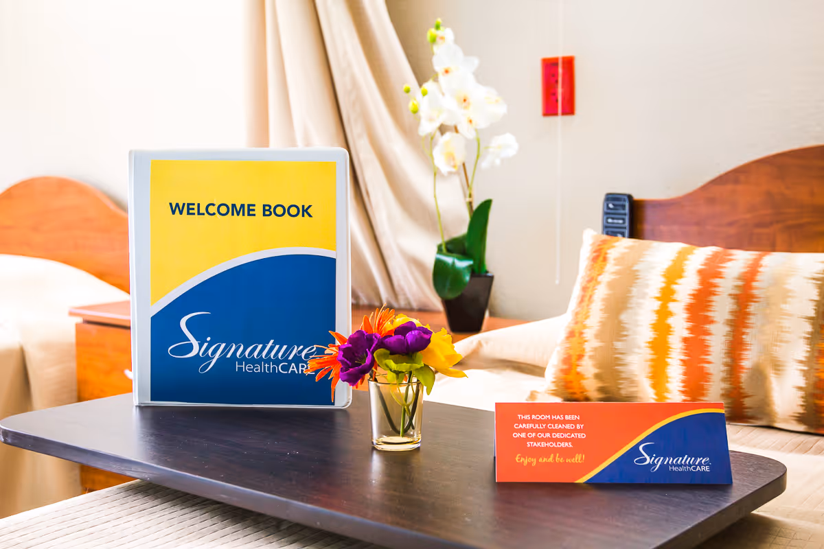 A bedside table in a bedroom with a welcome book from Signature HealthCARE, a small vase with colorful flowers, and a card indicating the room has been carefully cleaned. In the background, there is a bed with a patterned pillow and a potted orchid plant.