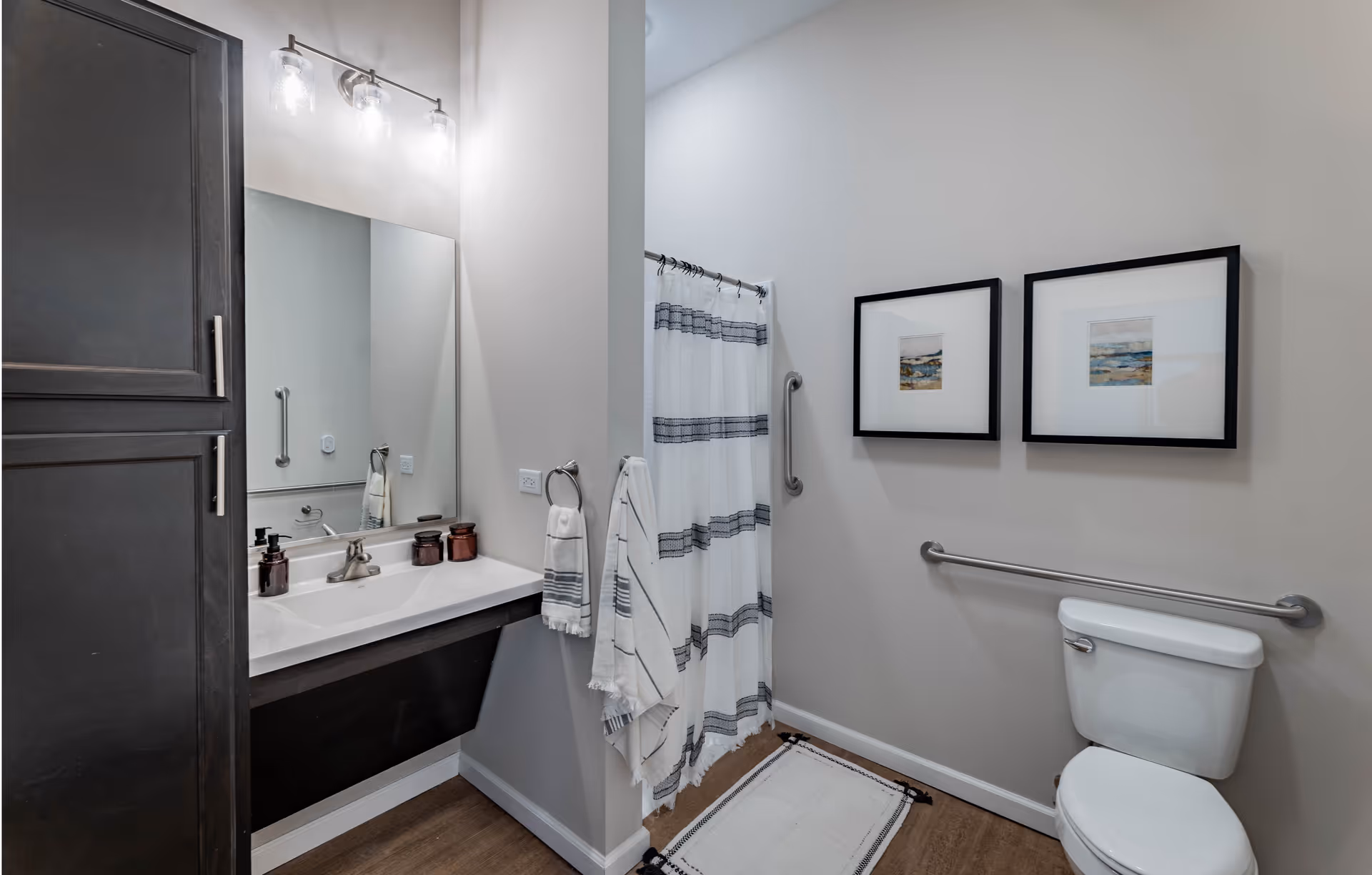 A modern bathroom with a white sink and dark cabinetry on the left, a large mirror above the sink with three light fixtures, a towel hanging on a ring, a shower with a white curtain featuring horizontal gray stripes, two framed pictures on the wall, a white toilet with a grab bar beside it, and a white rug on the wooden floor.
