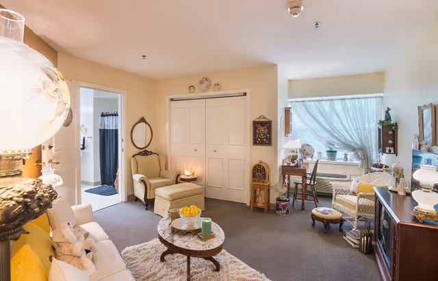 A cozy living room in a senior living facility with a white sofa adorned with yellow pillows and stuffed animals, a round marble coffee table with candles and a bowl of lemons, a beige armchair with an ottoman, a small wooden desk with a chair near a window with sheer curtains, and various decorative items including lamps, framed pictures, and plants.