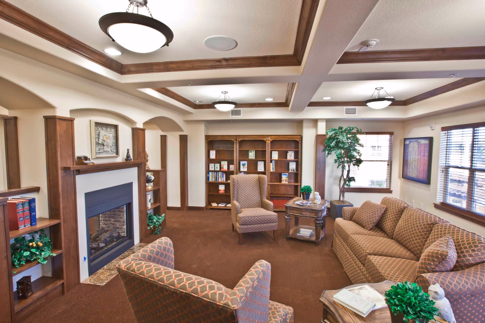 A cozy living room with patterned upholstered armchairs and a sofa arranged around a wooden coffee table. The room features a fireplace with a wooden mantel, built-in bookshelves filled with books and decorative items, and large windows with wooden blinds allowing natural light to enter. The ceiling has recessed lighting and wooden trim accents.