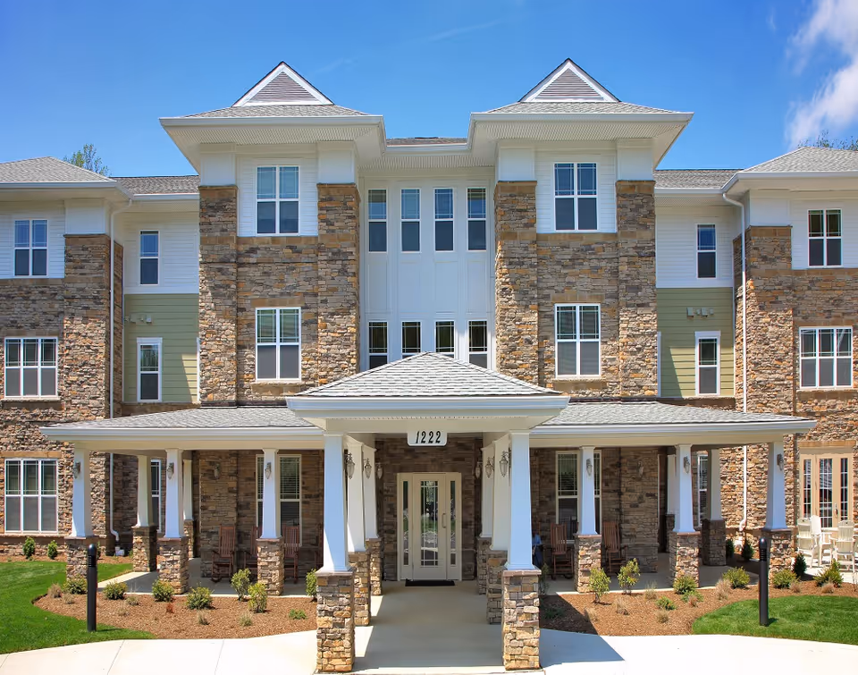 Front exterior view of a senior living facility named Perryman Station with a stone and siding facade, multiple windows, a covered entrance with white columns, and a well-maintained lawn and landscaping under a clear blue sky.