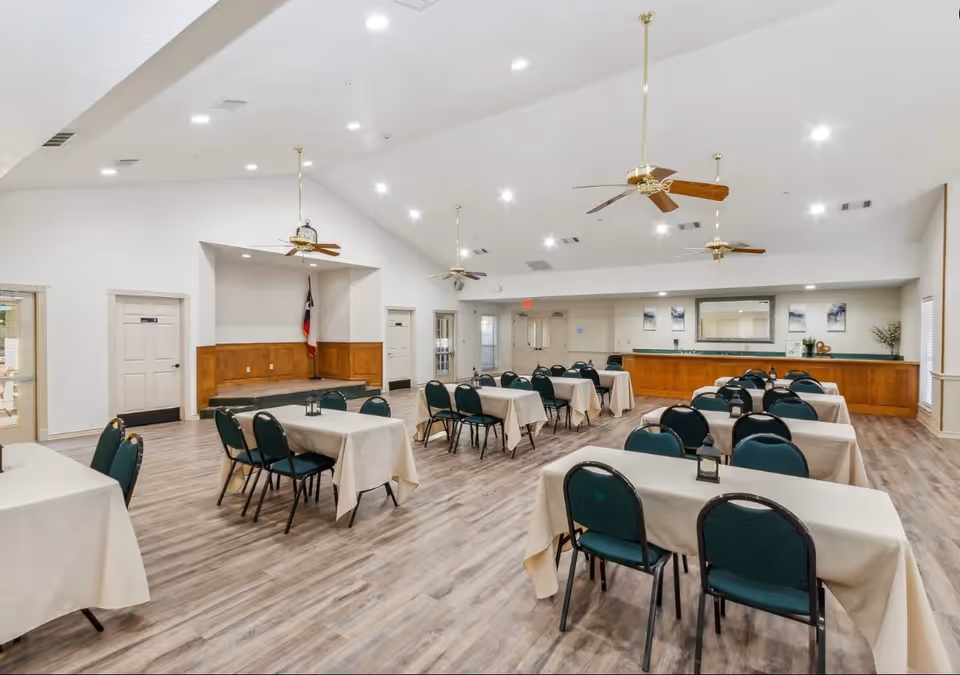 Large multipurpose dining/meeting room with rows of tables covered in tablecloths, green chairs, ceiling fans, and a serving counter.
