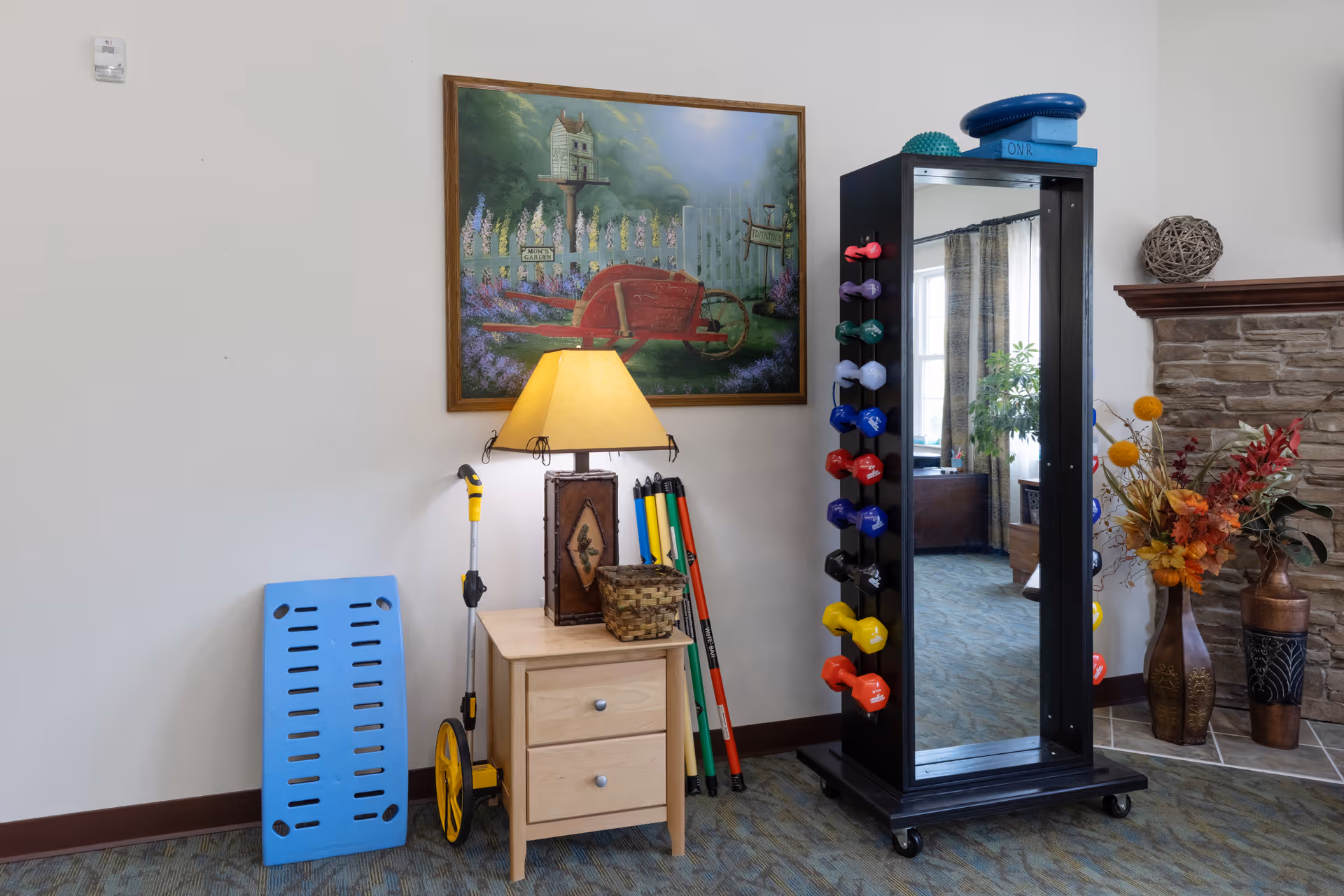 A bright interior common area featuring a small wooden side table with a lamp, a mirror-mounted dumbbell rack, exercise equipment, artwork, and decorative vases.