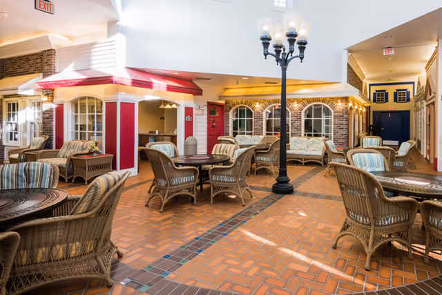 Indoor common area with wicker chairs and tables arranged on a brick-patterned floor. The space features a streetlamp-style light fixture in the center and has a cozy, village-like design with small storefront-style facades painted in red and yellow with windows and awnings. The area is well-lit with natural light coming from above.