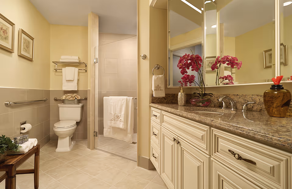Spacious, well-appointed bathroom with a granite-top vanity and large mirror, toilet, grab bars, and a glass-enclosed walk-in shower with decorative orchids on the counter.