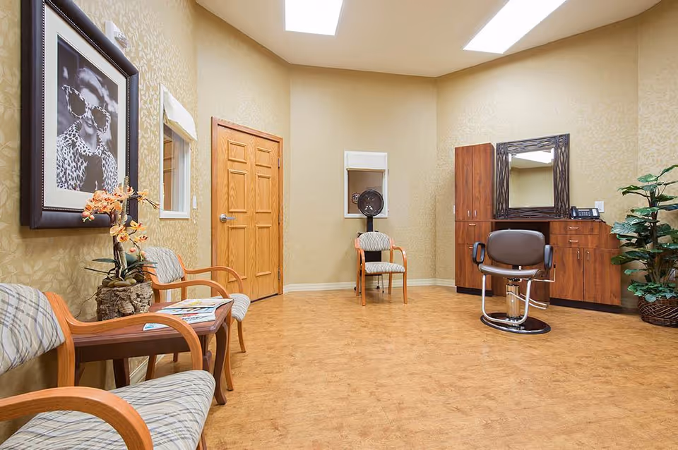 Interior of a salon room with wooden flooring and beige patterned walls. The room features a styling chair in front of a large mirror mounted on a wooden cabinet, two cushioned chairs with wooden arms, a small table with magazines and a flower arrangement, a hair dryer chair, a wooden door, and a potted plant in the corner.