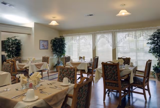 A dining room in a senior living facility with several tables covered in beige tablecloths, each set with white cups, napkins, and silverware. The room has large windows with sheer white curtains allowing natural light to fill the space. There are upholstered wooden chairs around the tables and potted plants placed near the walls.