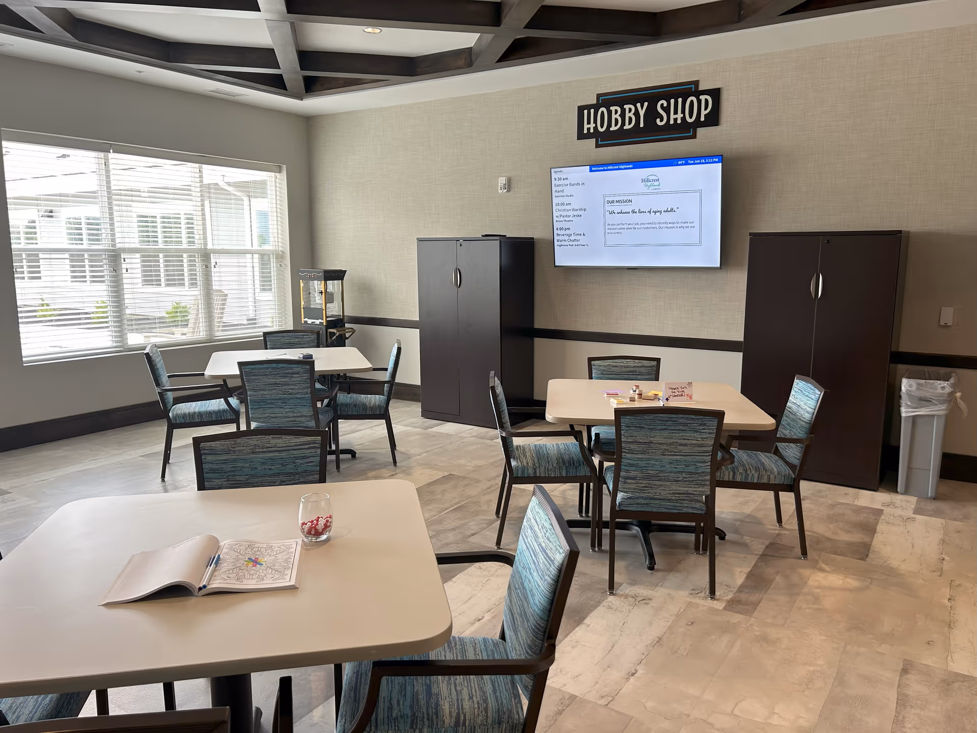 A well-lit hobby shop room with several tables and chairs arranged for group activities. There is a large window letting in natural light, two dark storage cabinets against the wall, and a flat screen TV mounted on the wall displaying information. A sign above the TV reads 'HOBBY SHOP'. On one table, there is an open coloring book and a glass with colored pencils.