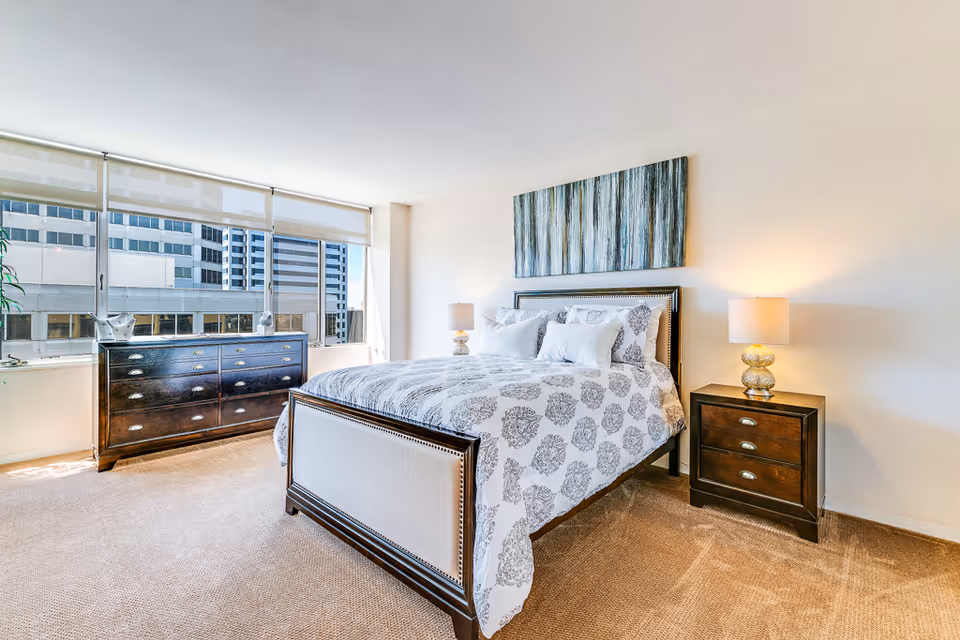 A bright and spacious bedroom with a large bed featuring white and gray floral bedding, two bedside tables with lamps, a dark wooden dresser, and large windows showing a cityscape outside.