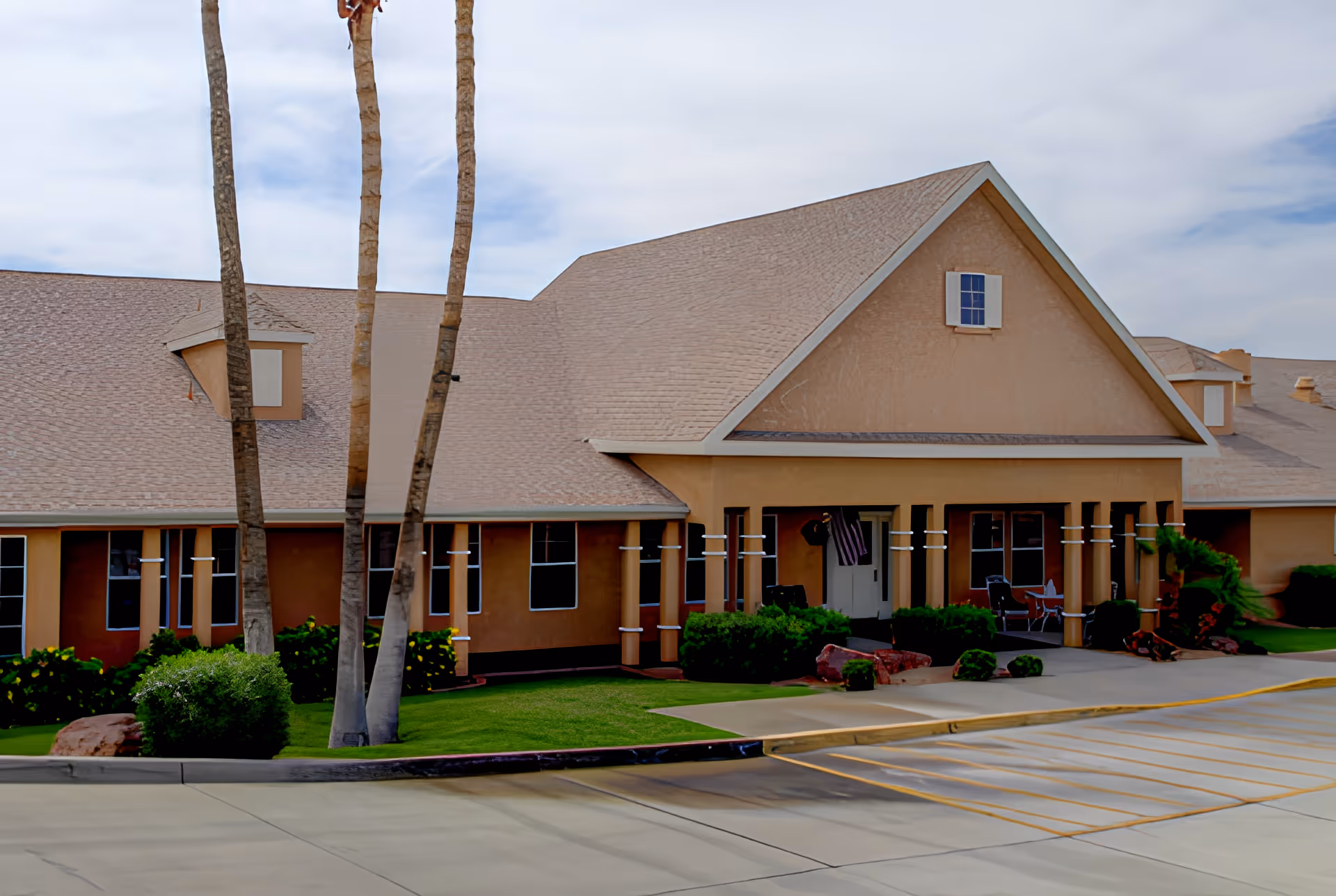 Exterior view of a single-story senior living facility building with a beige facade, multiple windows, and a sloped roof. There are palm trees and green bushes in front of the building, along with a paved parking area with marked parking spaces.