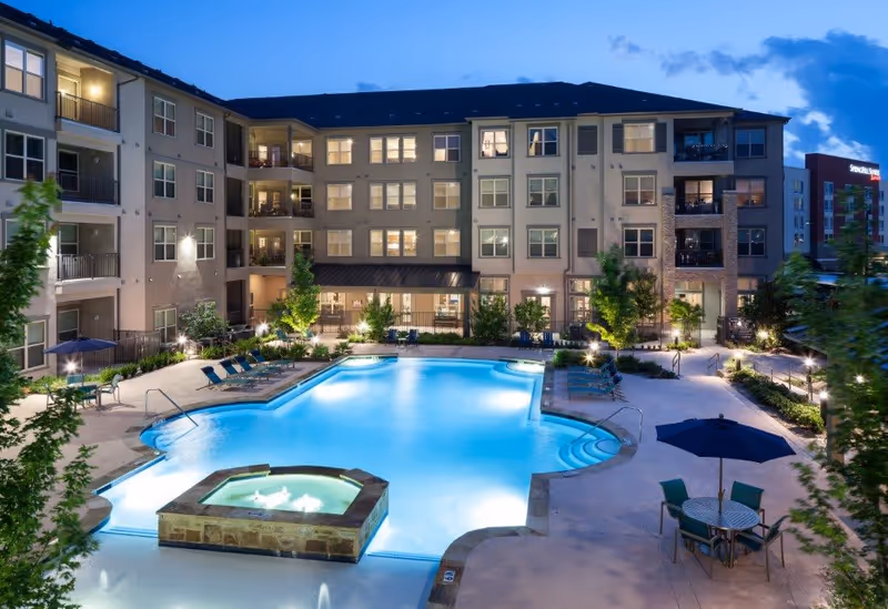 Evening view of a well-lit outdoor swimming pool area surrounded by a multi-story residential building with balconies. The pool area includes lounge chairs, a hot tub, tables with umbrellas, and landscaped greenery.