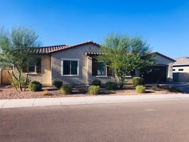Single-story house with a tiled roof, beige stucco exterior, and a two-car garage. The front yard features desert landscaping with small bushes and two medium-sized trees. The sky is clear and blue.