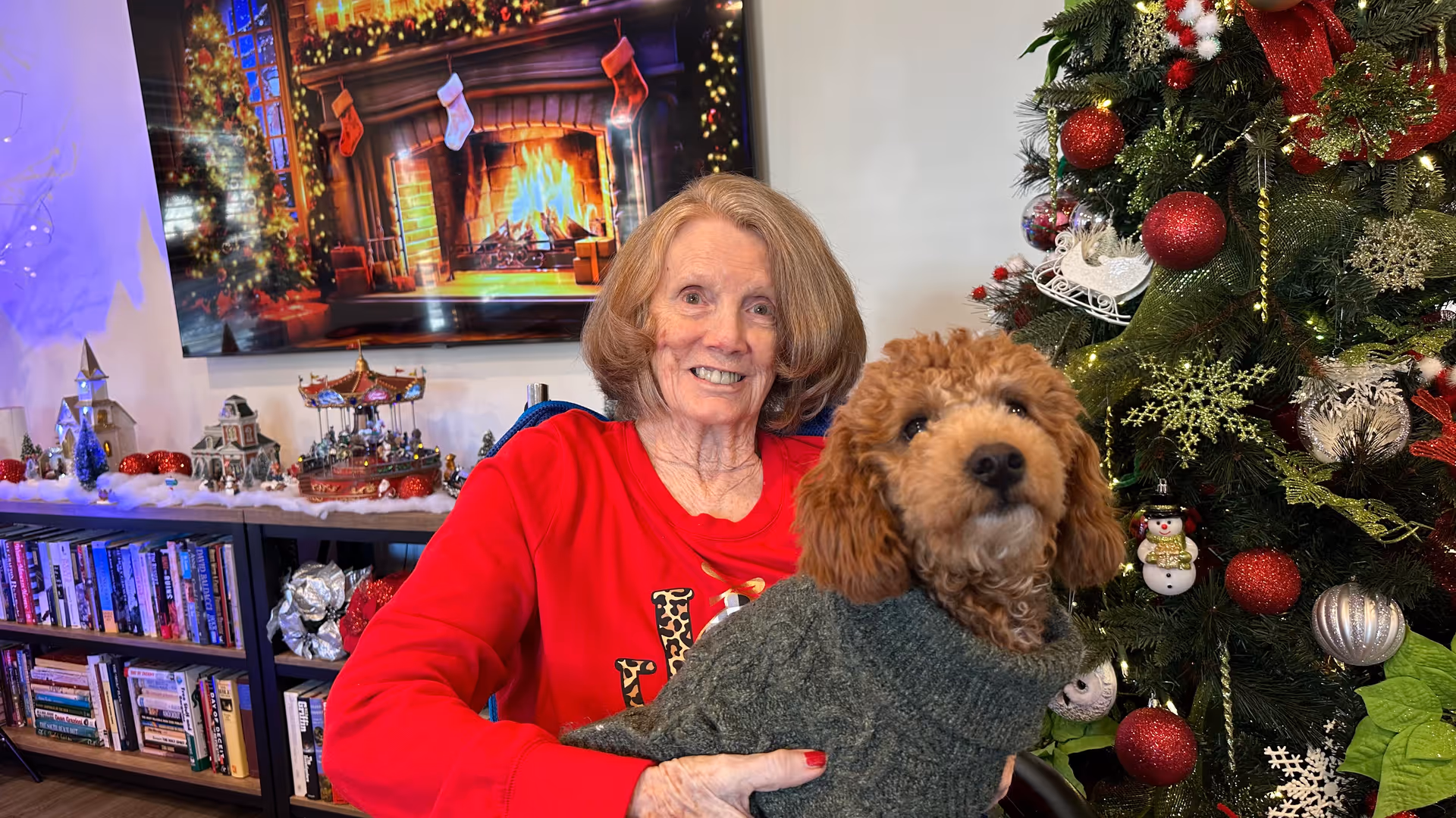 An older woman in a red sweater sits holding a small brown dog in a green sweater in a festively decorated room with a Christmas tree and a fireplace scene behind them.