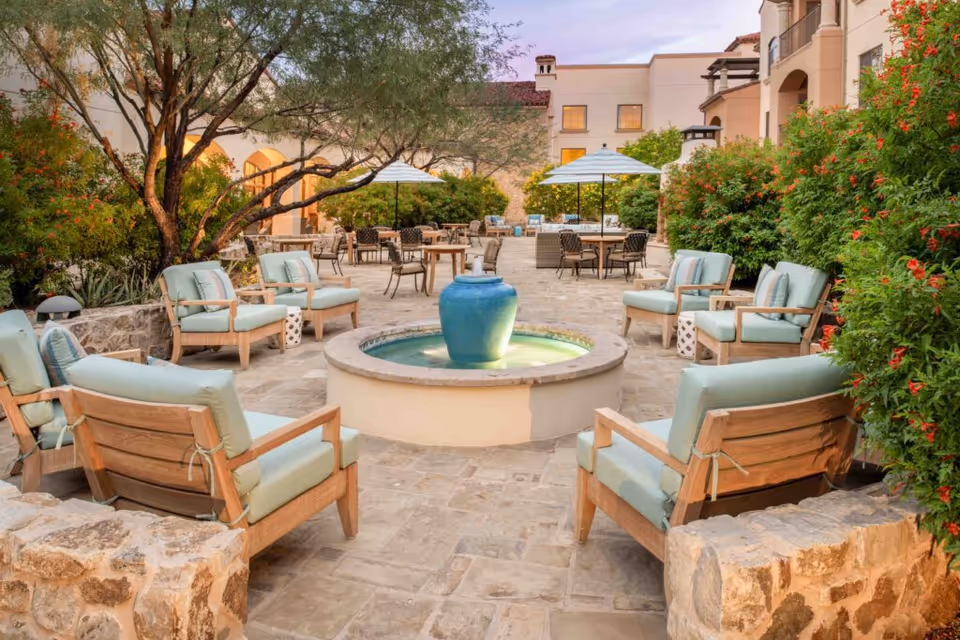 Outdoor courtyard area with cushioned wooden chairs arranged around a circular water fountain with a blue vase centerpiece. There are tables with umbrellas and additional seating in the background, surrounded by greenery and stone buildings under a clear sky.