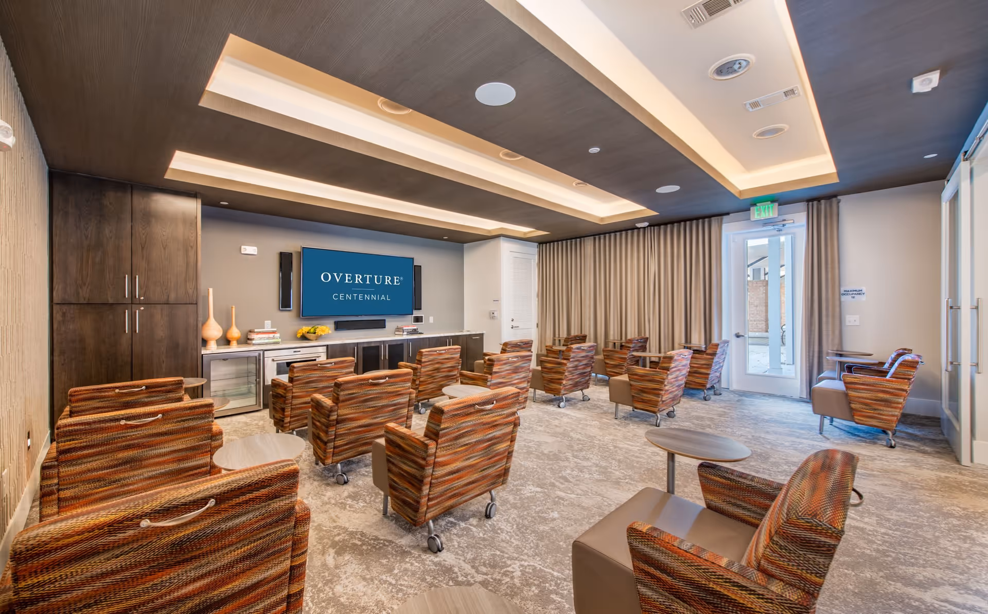 A modern meeting or media room with multiple striped armchairs on wheels arranged in rows facing a large flat-screen TV mounted on the wall displaying the text 'Overture Centennial'. The room features a dark wood ceiling with recessed lighting, a carpeted floor, a cabinet with decorative items and books, and a glass door with curtains on the right side.
