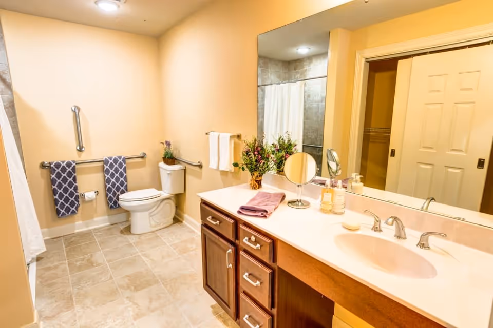 Bright and spacious bathroom with beige walls and tiled floor. The room features a white toilet with grab bars on the walls, two patterned blue towels hanging on a bar, and a white shower curtain. A wooden vanity with a white countertop holds a folded purple towel, a small round mirror, a vase with flowers, and soap dispensers. A large mirror is mounted above the sink, reflecting the opposite wall and a sliding door.